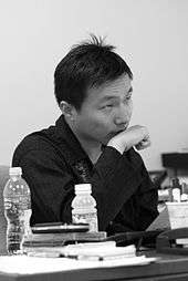 An Asian man in a dark shirt seated at a desk and looking right with his wrist held to his chin