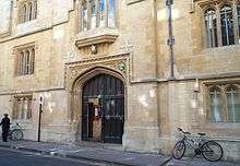 A stone building with a large archway of decorated stone; double doors (closed) in the archway; the left-hand door has a smaller inset door, which is open. Above the archway, an oriel window; some bicycles are leaning up against the walls to the side of the archway