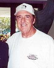 Man in his late 70s, smiling and wearing a white T-shirt and baseball cap.