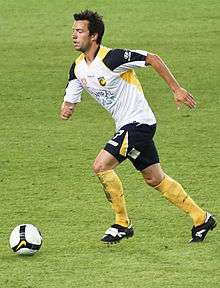 A man with dark hair in a white shirt and navy shorts running towards a football