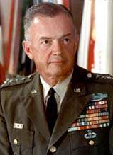 White-haired, clean-shaven man in dark-green ceremonial military uniform and tie. He has a lot of insignia on the left side of his chest and stars to indicate that he is a general.