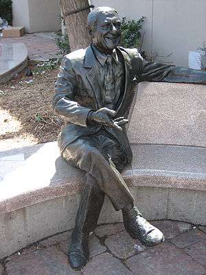 A statue seated on a curved bench, behind which is the trunk of a tree adjacent to the wall of a building.