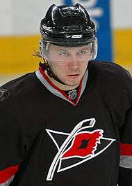 An ice hockey player standing directly in front of the camera. He is wearing a black helmet and uniform.