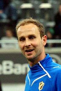 A photograph of a dark haired man with a blue training sweater, the man is seen from the side, he is smiling.