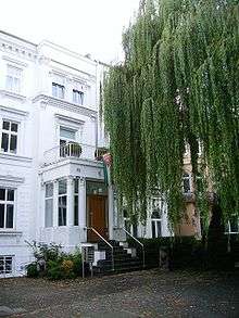 Parts of a white building with a tree in front. The entrance is on a higher level with columns.