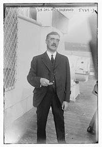 Three-quarter length photograph of a moustachioed man standing on the deck of a ship