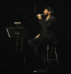 A bearded man in black clothing with his eyes closed. He is holding a microphone and sitting on a stool on a stage.