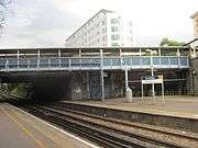 A canopied footbridge. Underneath the footbridge are a pair of railway tracks & platforms, approximately at a 55° angle. Behind the footbridge is a yellow brick wall. The wall has a hole in it to allow the railway tracks to go through into an area of green foliage. Behind the wall there is an apartment building. Underneath the footbridge there is a cycle rack & a blue, white & orange sign bearing the text "Kew Bridge" "Fare zone 3" "Alight here for The Royal Botanic Gardens". The weather is partially cloudy.