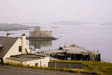  A white building sits above a concrete pier. Offshore there is a small island on which there is a building with grey featureless walls. Various other islets can be seen in the background through the mist.