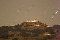 Time-lapse photograph of white buildings on top of a mountain at night with stars streaking across the sky.