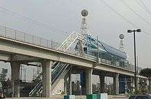 An elevated train station and two train tracks. The station has a blue roof and is made of white metal.