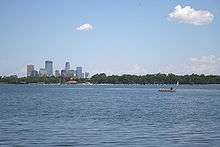 Light clouds sit in a blue sky over a wavy lake; two people fish from a boat on the right, while Minneapolis's downtown skyline jumps above the treeline to the left.