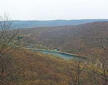 A narrow lake with buildings on the far side in woods just starting to green with the spring, seen down the slope of a mountain