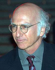 A bald man with white hair around his ears. He is wearing a black suit, blue shirt, glasses and a red tie.