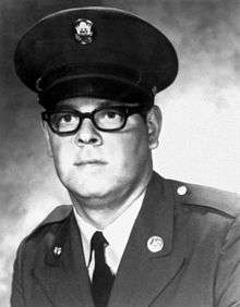 Head and shoulders of a young man with thick-framed glasses in military uniform. He is wearing a peaked cap and a military jacket with a "US" pin on the lapel over a shirt and dark tie