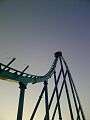 A photograph of a roller coaster train approaching a drop. The track is illuminated in green and blue under a dusk sky.