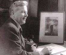 A black and white photograph of a smiling man seated at a desk. He is holding a pen, with a jotter open in front of him and a photograph on the desk.