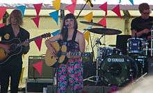 Mitchell and her two backing band members are shown in three-quarter shot. She stands in the middle facing forward while playing a guitar and singing into a microphone. Her right had strums the strings while her left is low on the neck. She wears a floral one-piece with pink and red flowers and green leaves. To her right is a man playing a guitar and to her right is a drummer at his kit, partly out of shot. About them is other stage equipment and coloured triangular flags.