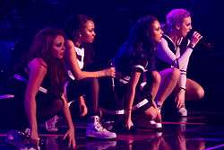 Four women on a stage with microphones in their hands. All four women are in a bending position and are wearing black and white outfits as bright purple and dark blue stage lighting shines upon them.