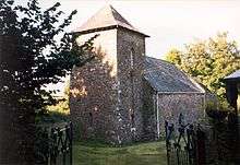 A stone church with a slate roof seen from the southwest. Nearest is a relatively large tower with a pyramidal roof, beyond which is body of the church