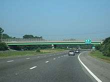 Ground-level view of a three lanes of a divided freeway; a large green and gray overpass bridge and a green exit sign are visible in the distance.