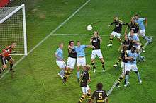 An action shot of two competing football teams trying to win a flying ball in the penalty area of one of the teams, the ball is heading for the head of the attacking teams captain. The players of the attacking team are wearing sky-blue shirts, white shorts and sky-blue socks. The field players of the defending team are wearing black shirts, white shorts and black socks. The goalkeeper for the defending team is wearing an orange shirt, black shorts and black socks.