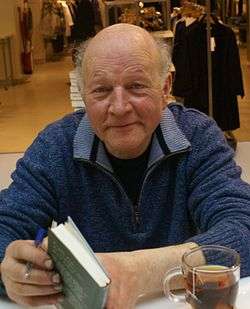 Maarten 't Hart sitting behind a table and holding a book