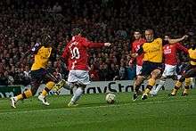 Wayne Rooney (in red) is shaping to kick the ball with an Arsenal player (in yellow) on either side of him. Cristiano Ronaldo, Carlos Tevez (both Manchester United) and Kolo Touré (Arsenal) are looking on in the background.