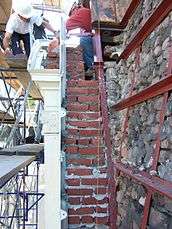 Cross-section of brick masonry exterior wall with cast iron cladding. Workers set new brick courses against the existing masonry back-up.