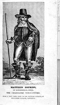 Black and white image of Hopkins. He holds a stick in one hand and has the other placed on his hip, and wears a large hat and wide boots.