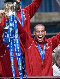 A man with very short hair is wearing a red jacket. He is holding aloft a trophy.