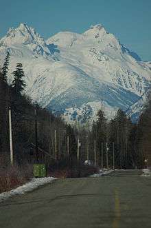 A multi-peak mountain raising above trees and a paved road