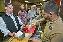 Several people lined up alongside a bar in a small area with fake wood paneling. Behind the bar, on the right, a bartender pours a drink for one of them.