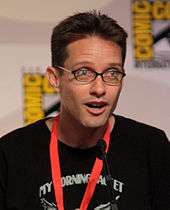 A man with dark brown hair and glasses leaning slightly forward, and speaking into a microphone.