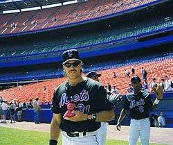 A man in a black baseball jersey and cap with "Mets" across the front in blue script stands on a baseball field.