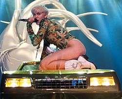 A short-haired blonde woman dressed in a marijuana-themed leotard is kneeling on a green sport utility vehicle with a microphone held to her mouth.