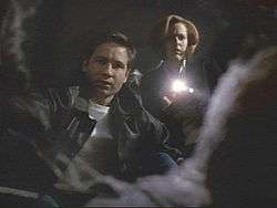 A man in a leather jacket and a woman with a flashlight look into a hole that contains a corpse.