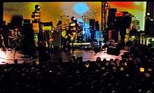 A five-piece rock band performing onstage against a psychedelic style yellow backdrop. Audience members facing the stage are visible in the foreground; various equipment including amplifiers, effect pedals, monitors and guitars are visible in the background.