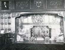 A black and white photograph of a fireplace with ornate mantel set in wood-paneled wall with similarly ornate carvings above it