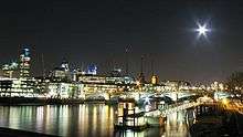 Nighttime photograph of illuminated buildings and bridge, with their lights reflected in the water.