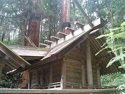 A simple wooden building with a railed veranda horizontal logs on the roof ridge and forked finials. From the front another roof runs over the steps to the veranda and connects to another building.