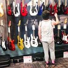 A guy with a white shirt and gray sweatpants is standing across a variety of electric guitars, with black guitar cases on the floor.