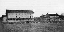 Black and white image of two structures linked by a covered verandah, the left building is three stories tall and much bigger than the house at right