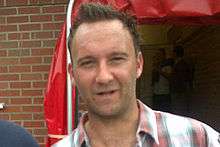 A man with an open multicoloured collared shirt smiles into the camera.