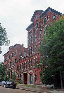 A large brick building, five stories on the right, three in the middle and four on the right, seen from across a cobblestone street. On the pediment atop the left, the words "Hinckel Brewing Company" can be seen.