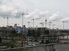 At least a dozen cranes rise over buildings under construction. In the foreground, cars are driving by on existing streets, and a city skyline rises in the background.