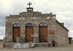 Photograph of Our Lady, Queen of Heaven Church, a small, one-story, stone church surmounted by a bell and wooden cross