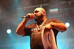 A bearded man wearing sunglasses, a chain, and a t-shirt reading 'CHABOS', rapping on-stage.