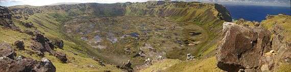 Colour photo of circular pond strewn marsh surrounded by steep cliff topped slopes - the sea is visible through a gap in the cliffs on the right