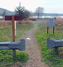 A footpath through low vegetation, starting from a gap in metal guardrails in the foreground, leading to some railroad tracks in front of water in the background. The rear of a sign is on the left, in front of the guardrails, and further back on the right is a large sign in blue type on white explaining that this path is for access to northbound service only and that southbound travelers should go a thousand feet up the road